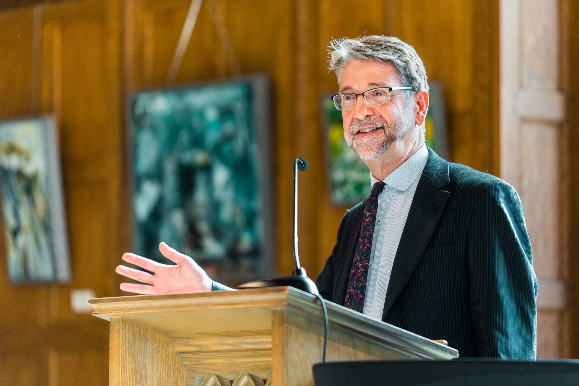 A man stands at a microphone, paintings behind him