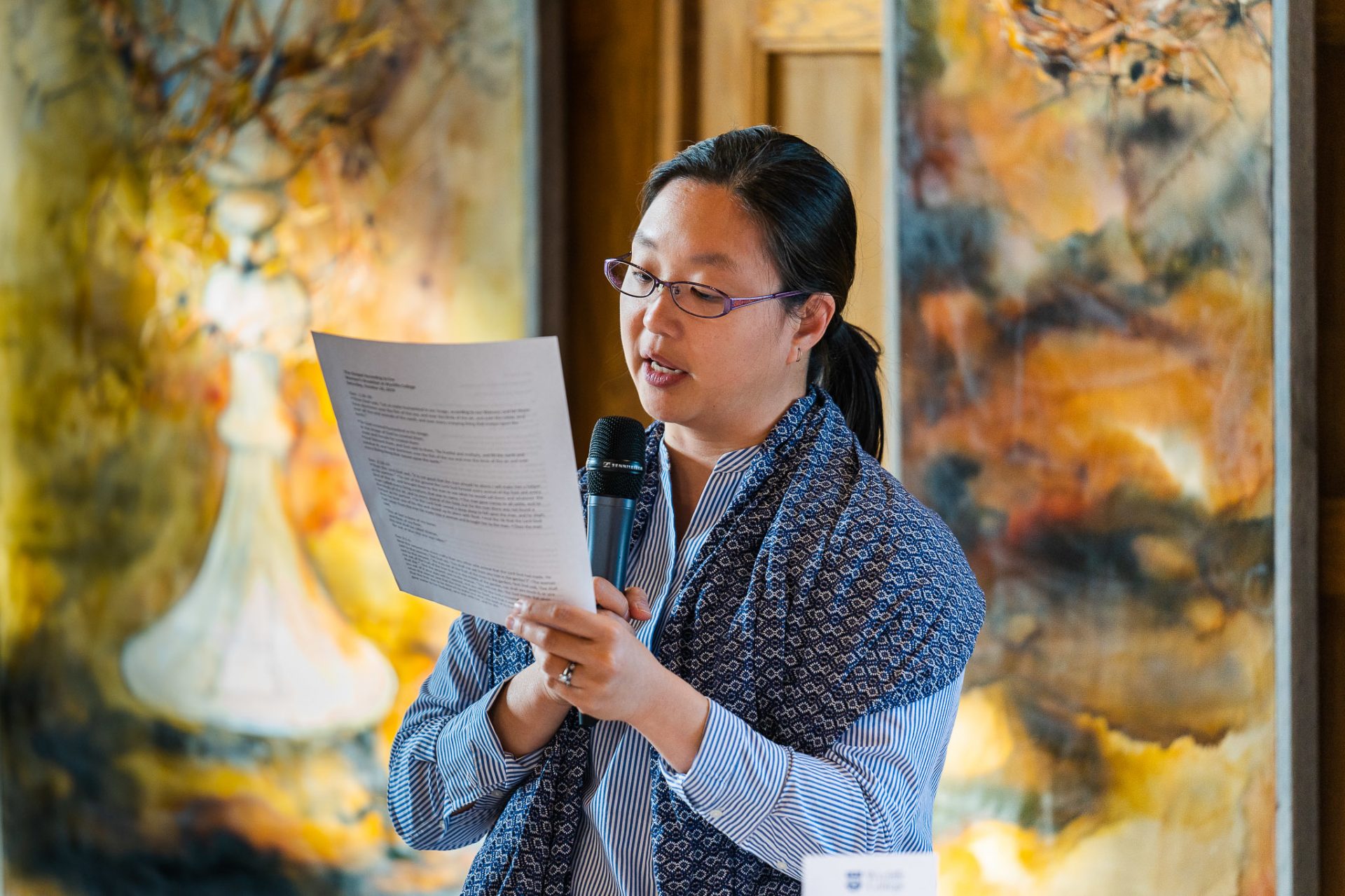 A woman stands in front of paintings, reading a piece of paper aloud into a microphone