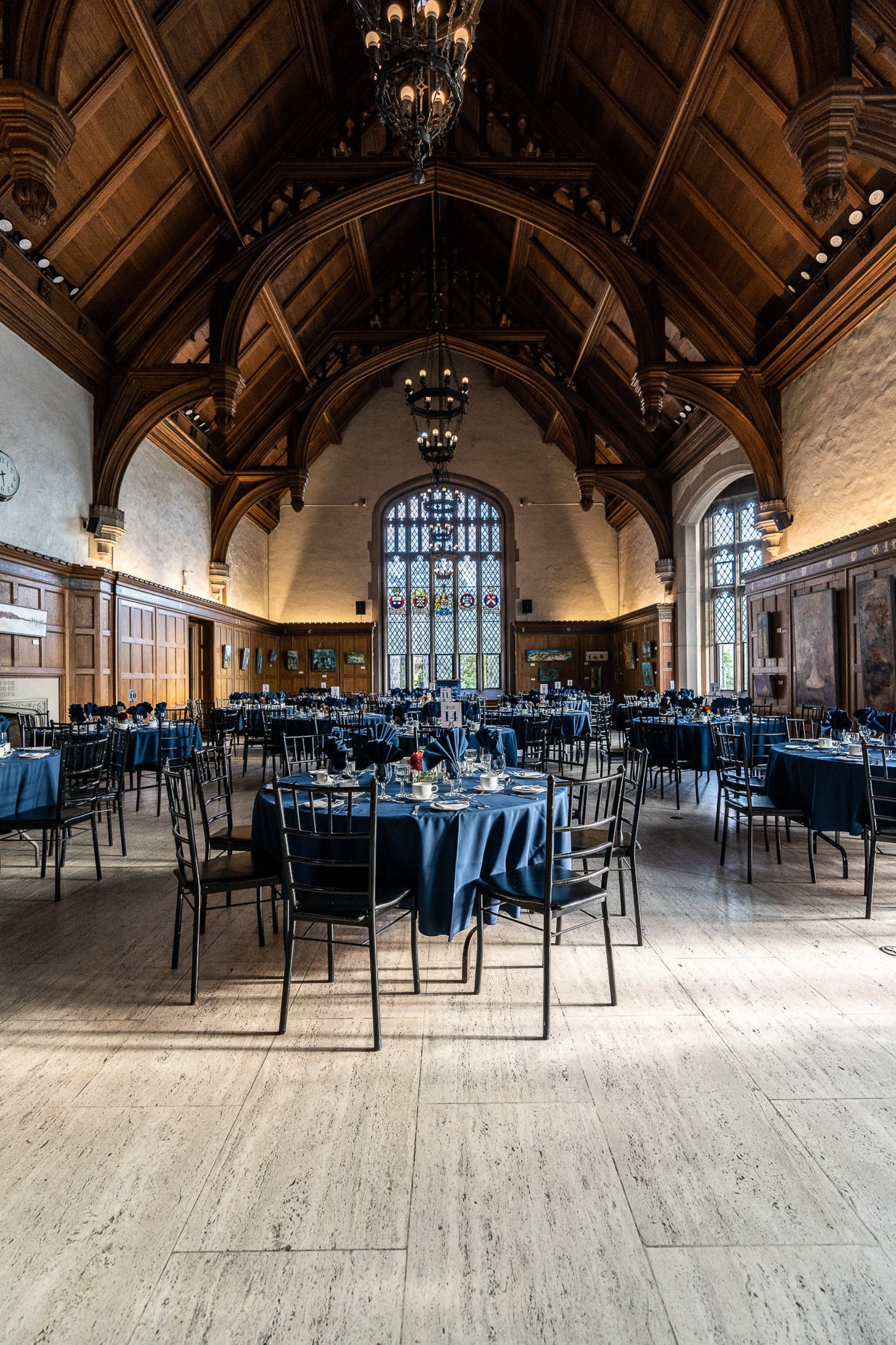 A group of tables set up for dinner in a large dining hall, light pouring through twenty-foot windows