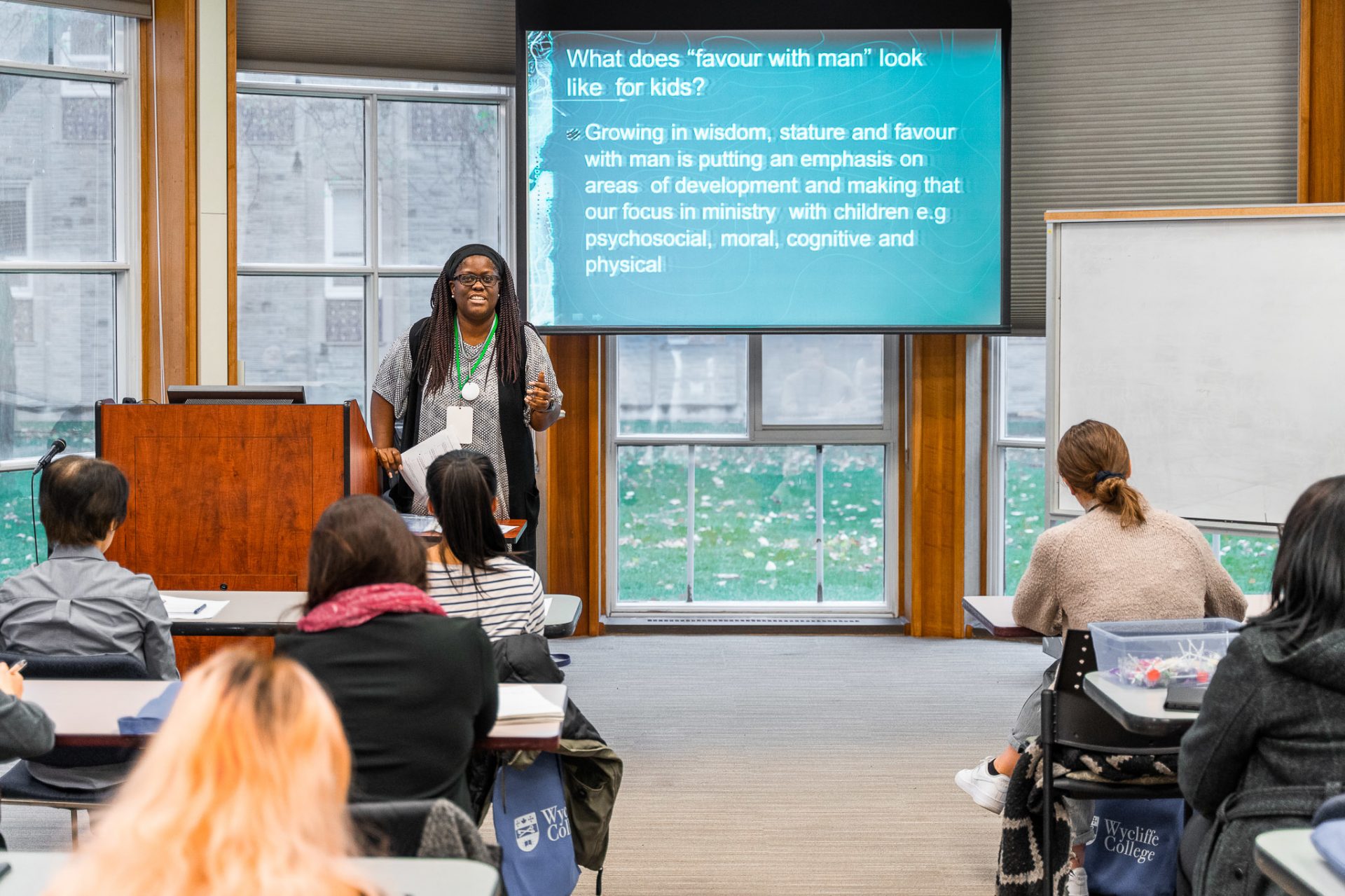 A woman teaching at the front of a classroom surrounded by glass windows.