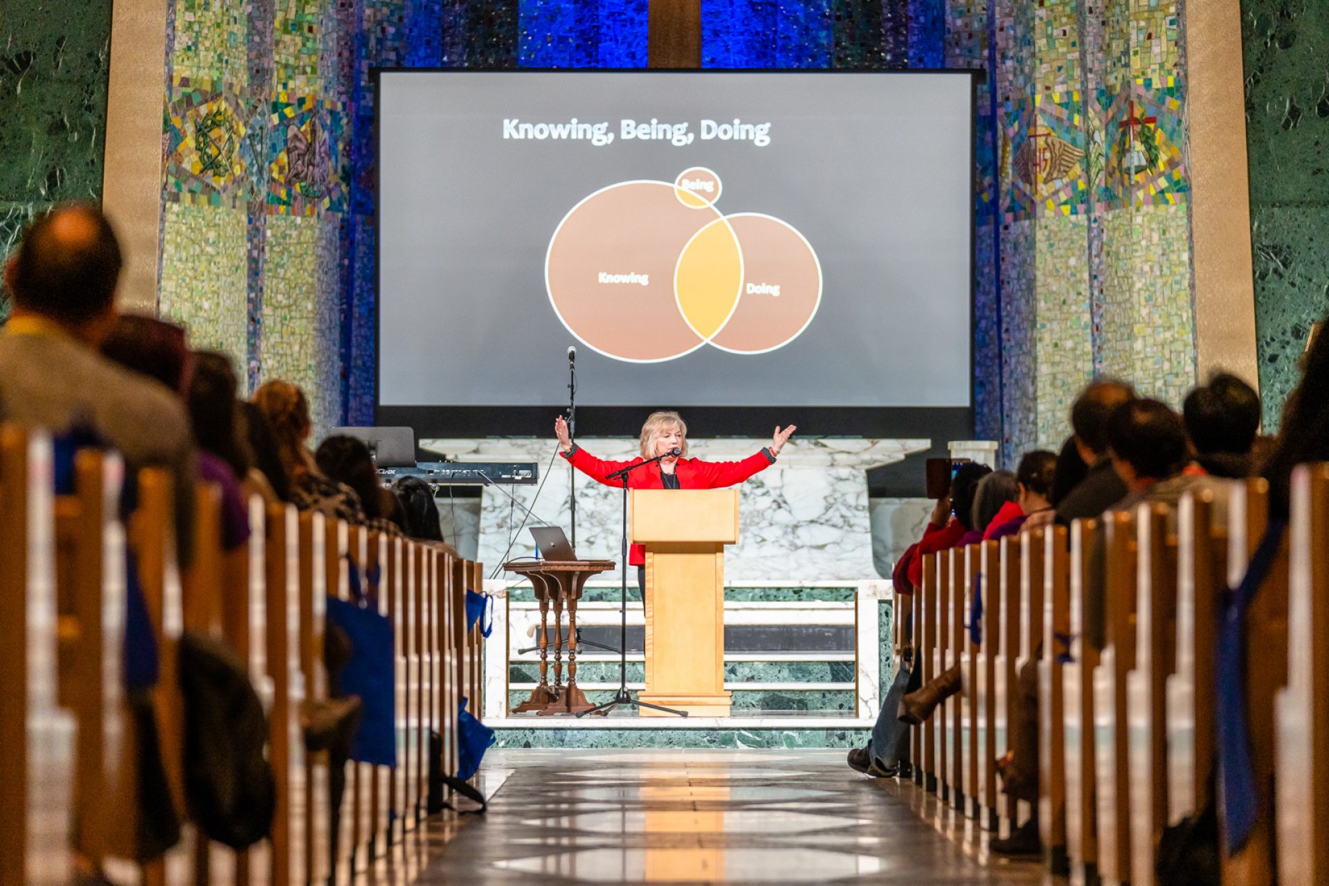 A woman lifts her hands up at a pulpit. The screen behind her displays a Venn diagram.