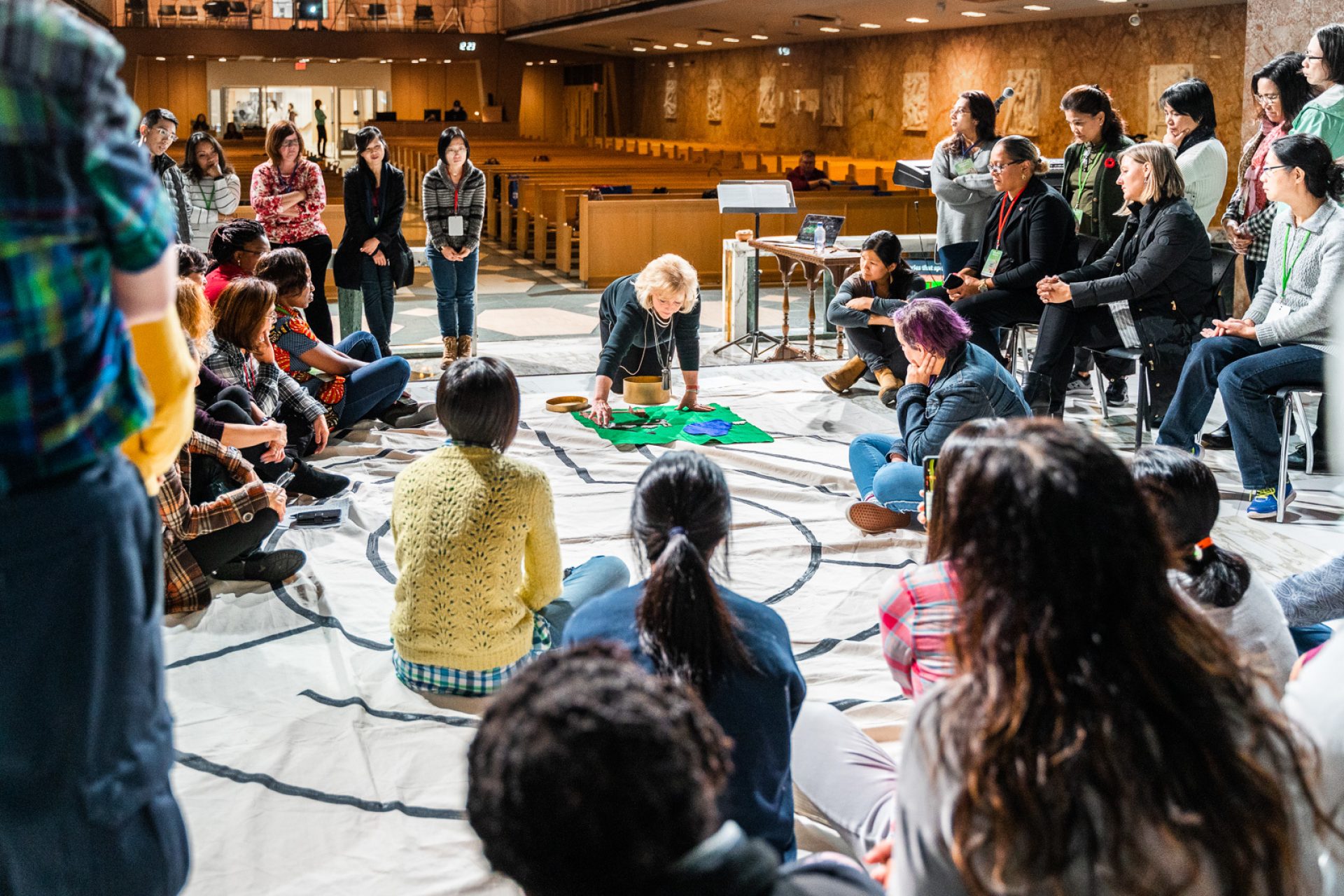 A woman leads a classroom in an illustration. They all sit in a circle around her on the floor as the lays out building blocks.