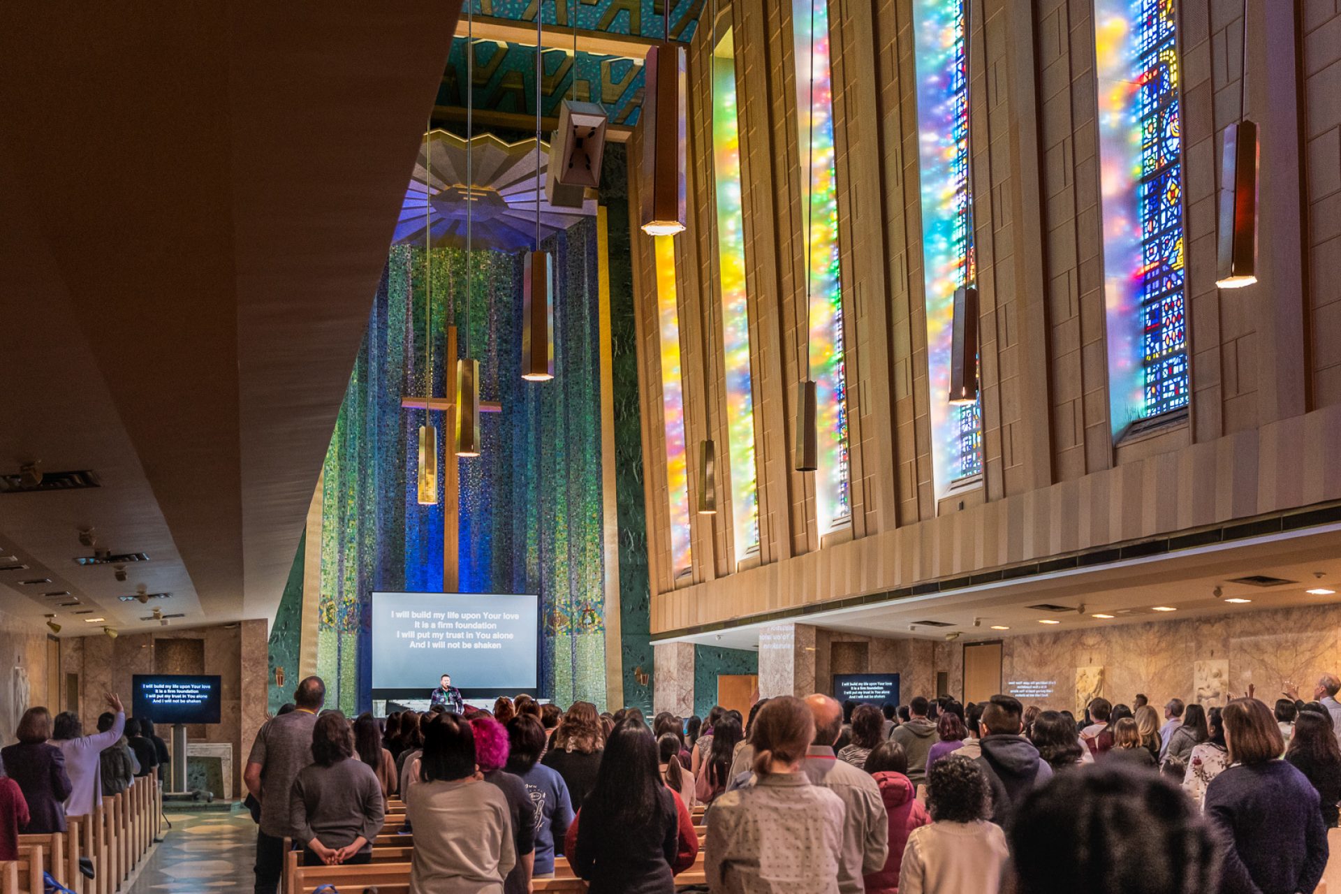 Light pours through stained-glass windows in a large chapel as a man leads a large congregation in worship